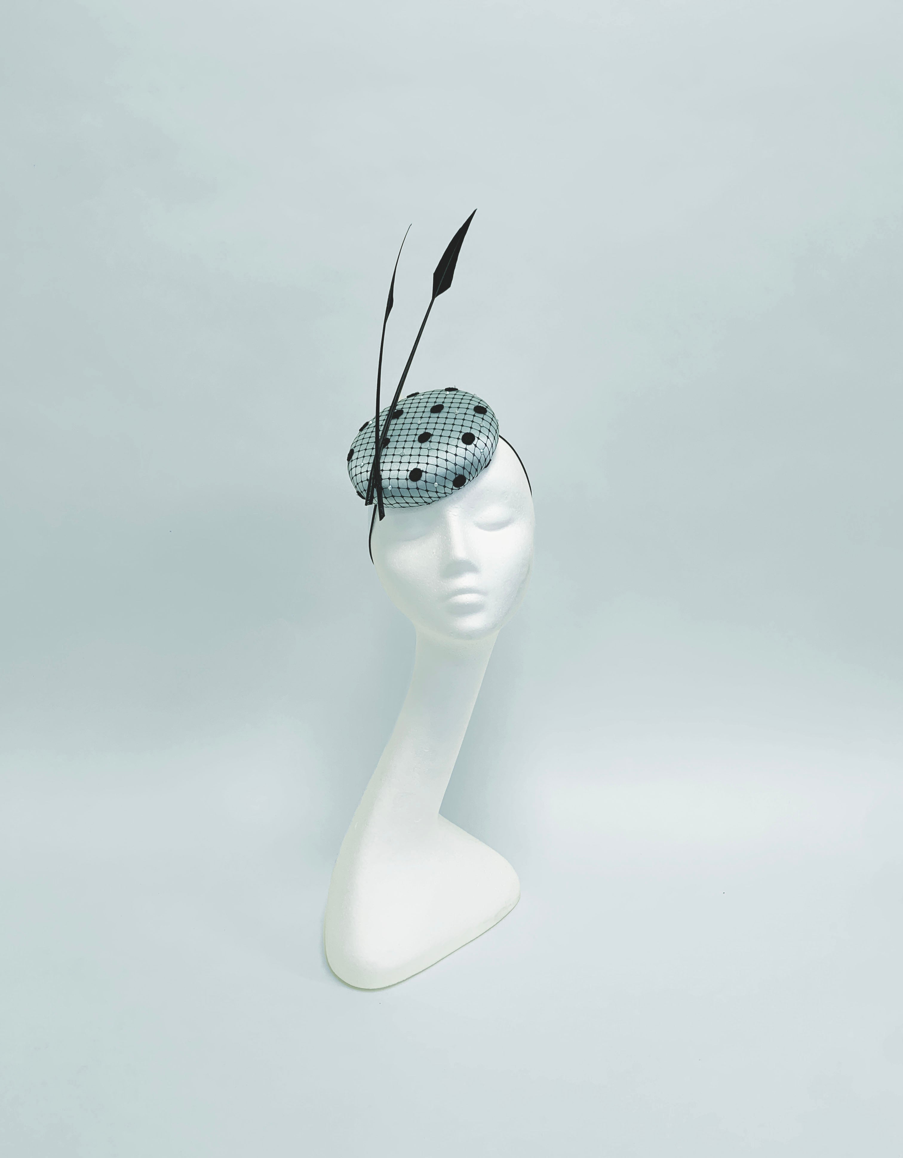 Black and ice blue polka dot pillbox hat with feather on a mannequin head against a light gray background