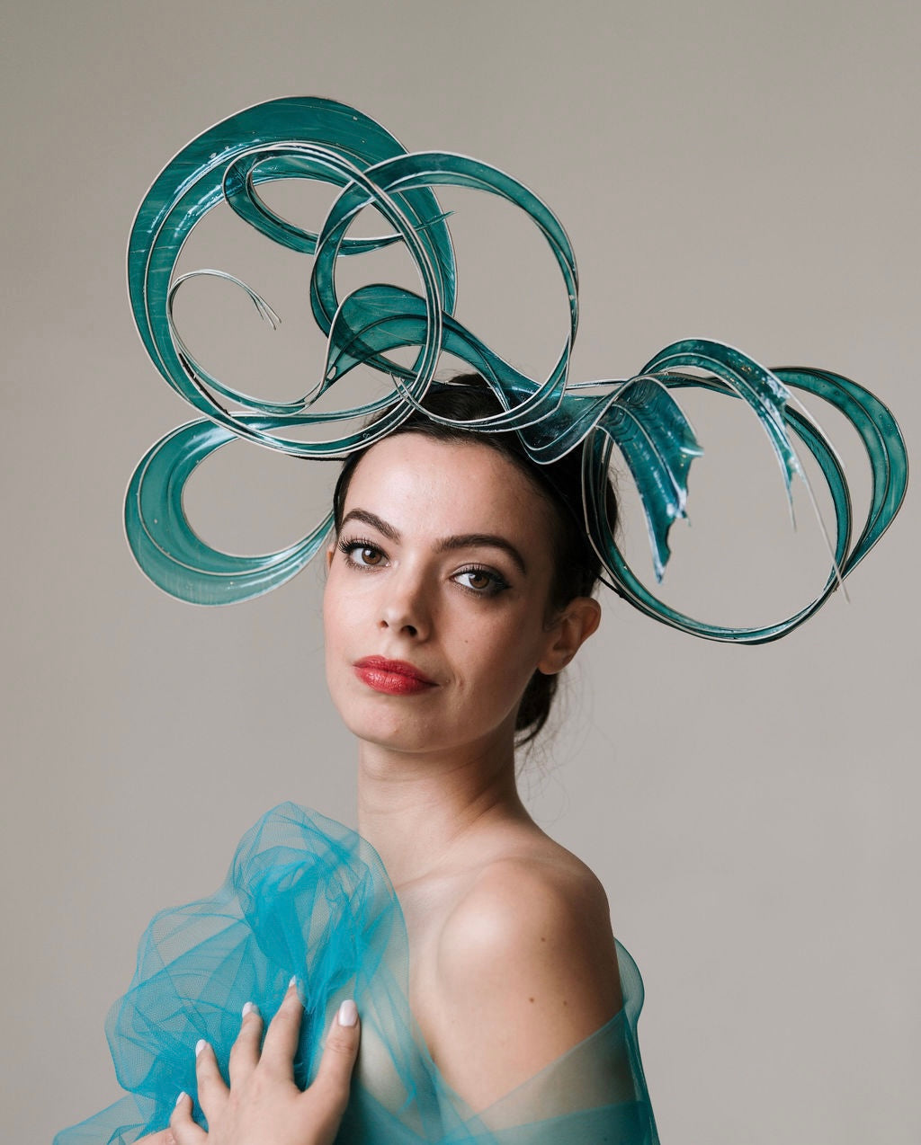 Woman wearing a large, decorative teal headpiece against a neutral background